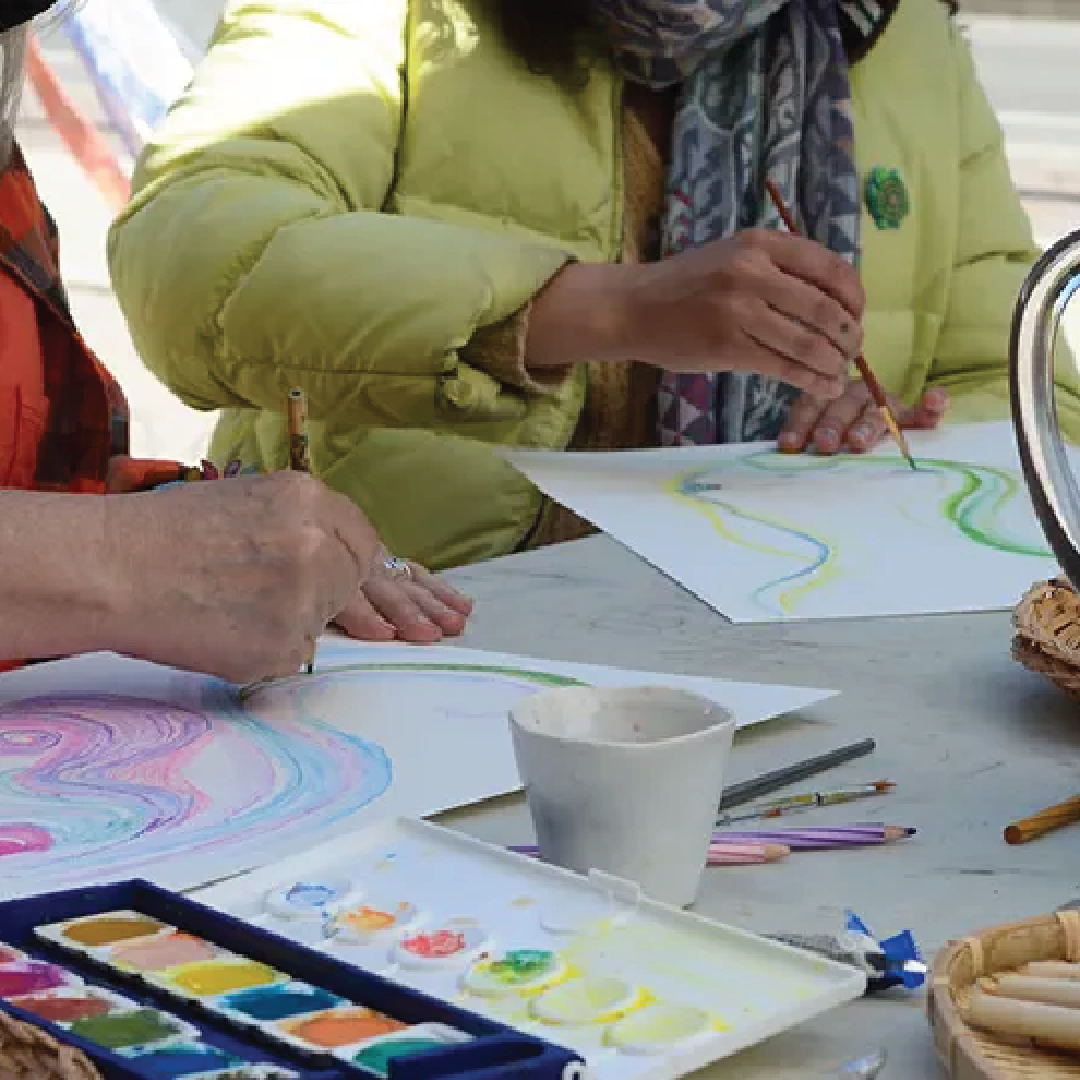 close-up of two people sitting at a table. one is drawing, the other is painting 