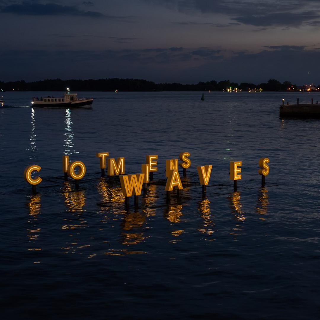 Floating artwork at night on water with big letters reads It Comes In Waves.