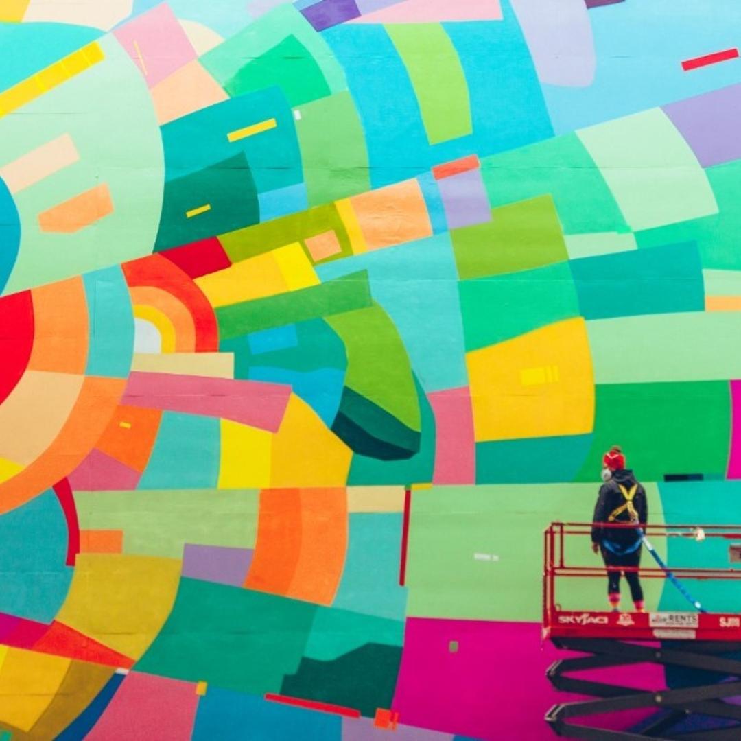 an artist on a scaffold painting a bright and colourful mural on a building