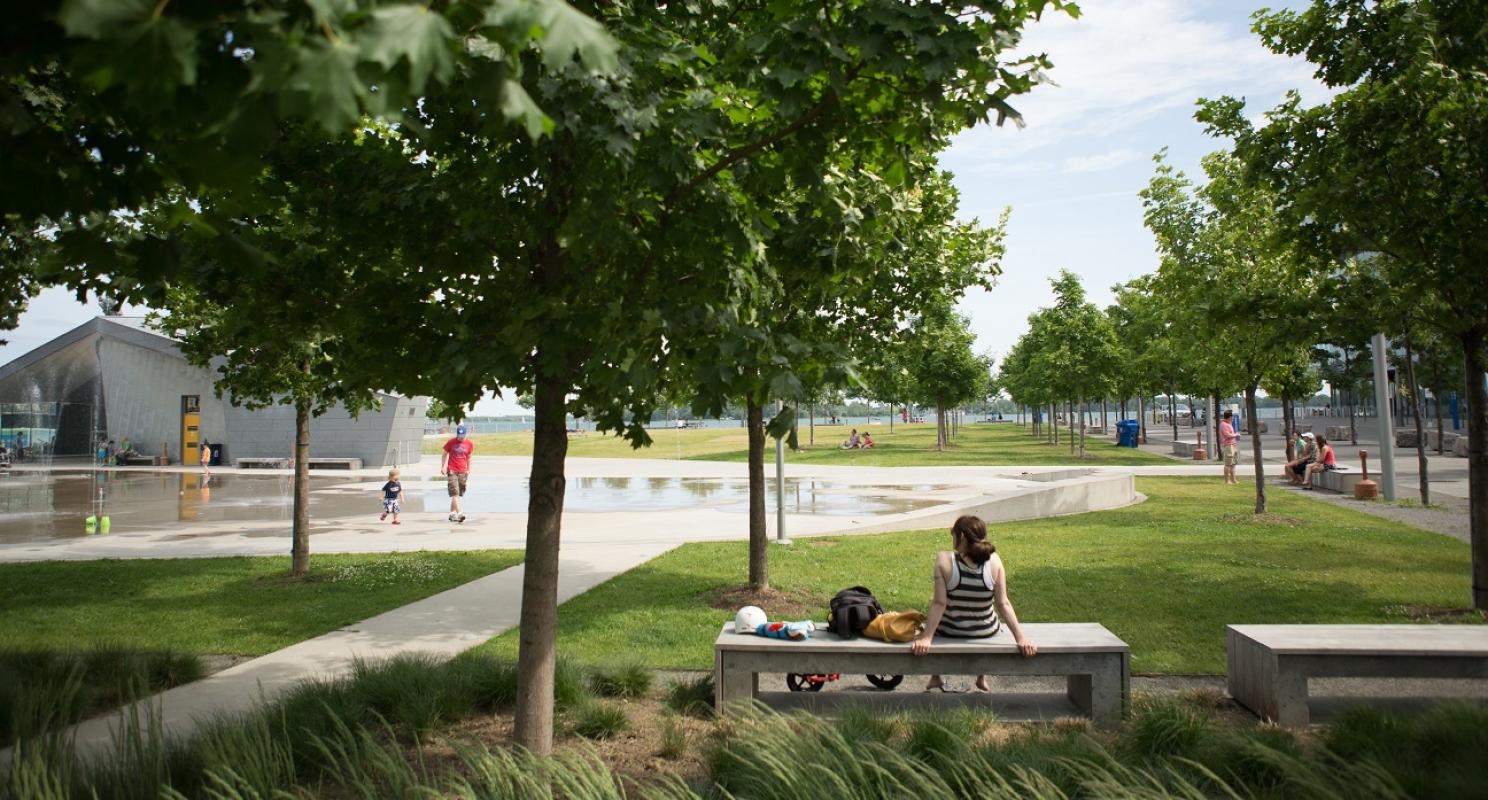 people sitting on a park bench enjoying the shaded green space next to trees, a pavilion and splashpad
