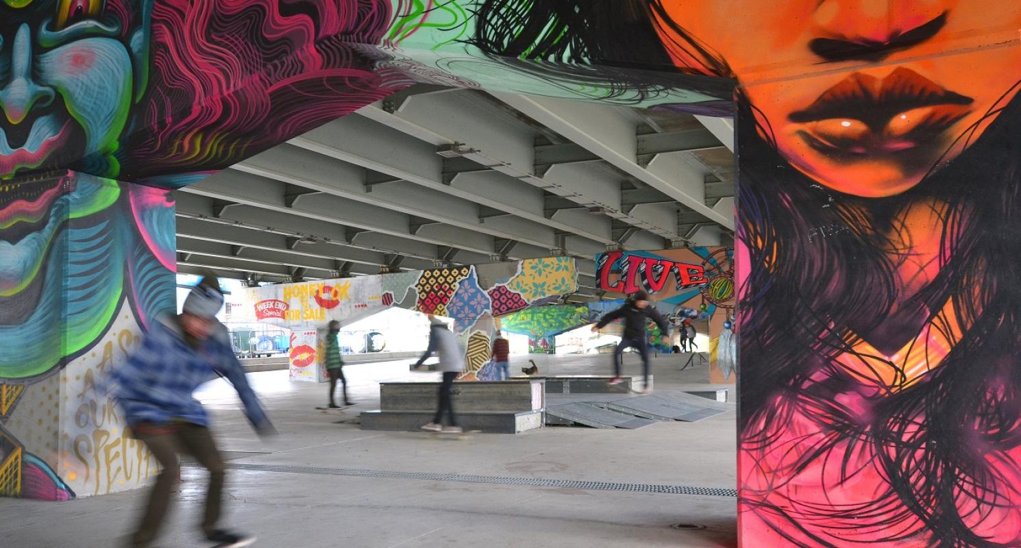 a group of skateboarders in an open space under an overpass with beautiful mural