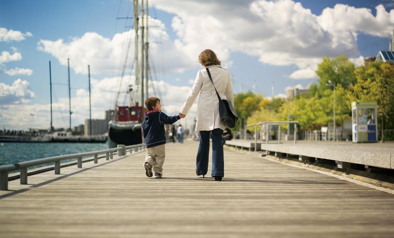 a mother and son holding hands walking along the water's edge promenade on a sunny day