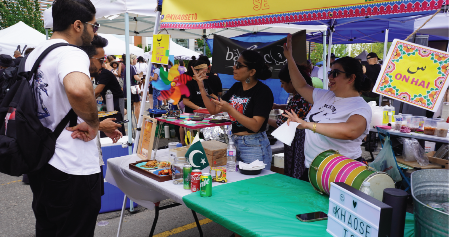 Two people at a vendor booth at a busy outdoor food market. 