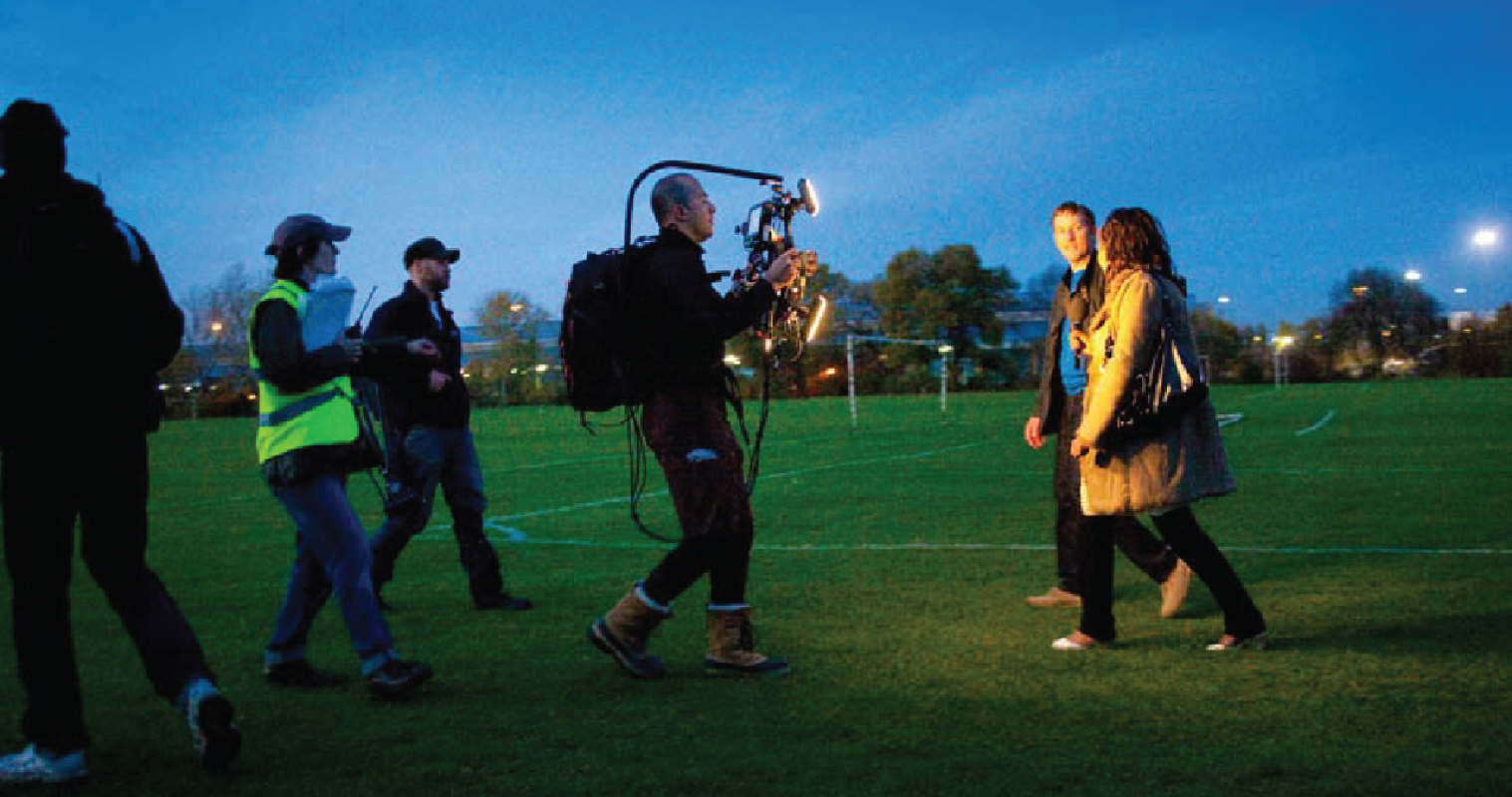 A camera crew filming two people walking on grass at sunrise.