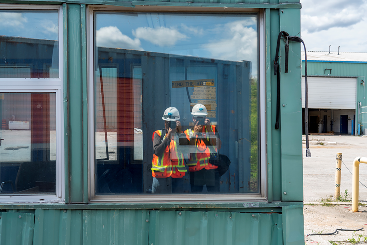 Two photographers in construction safety equipment taking a picture of themselves in a window.