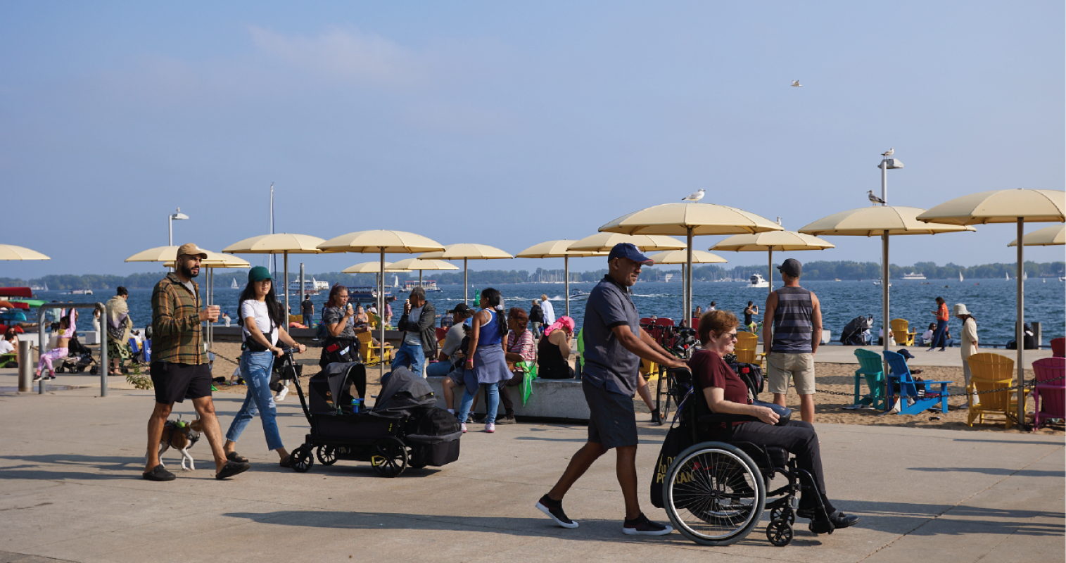 People at a beach on a summer day. One person is using a wheelchair, another is pushing a stroller. 