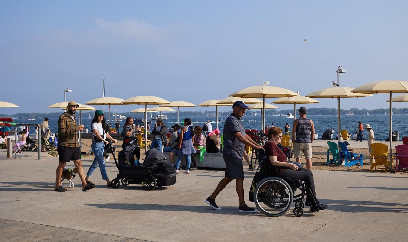 People at an urban beach on a summer day. One person is using a wheelchair, another is pushing a stroller.