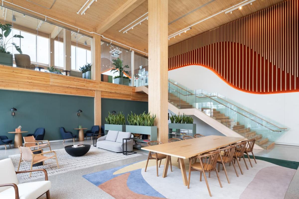 Modern office lobby with wooden accents and a variety of seating areas, featuring a large wooden table, sofas, and armchairs. The space includes lush green plants and a staircase with glass railings leading to an upper level. Decorative elements include a hanging wooden slat feature and green wall panels.