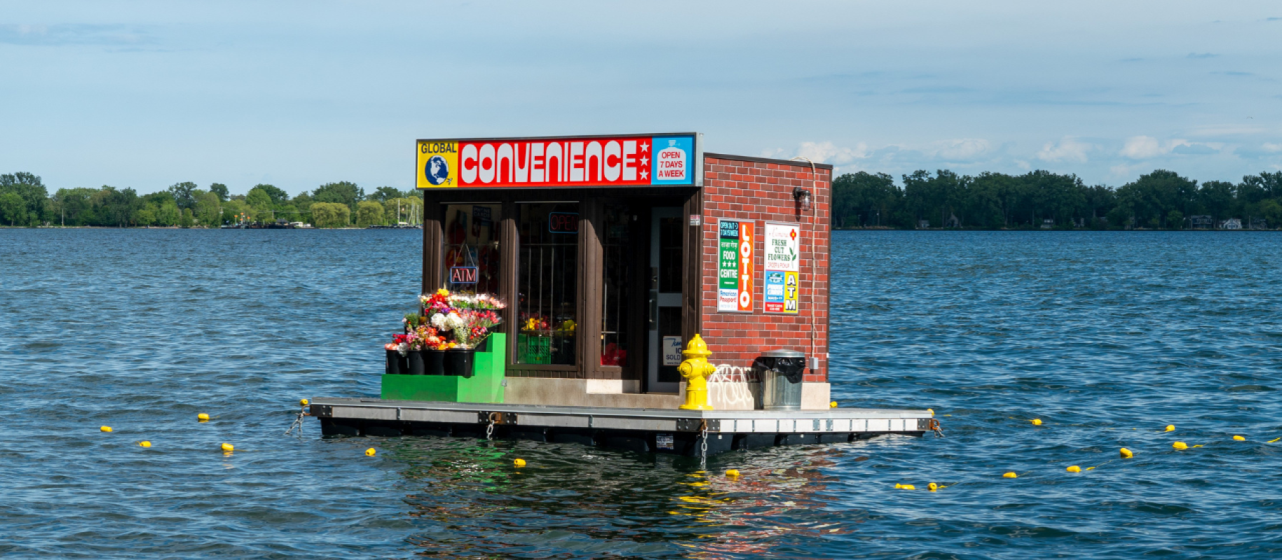 A floating convenience store artwork stocked with flowers and everyday items sits on the water in daylight, featuring a bright “Convenience” sign, an “ATM” sign, and a yellow fire hydrant on the platform.