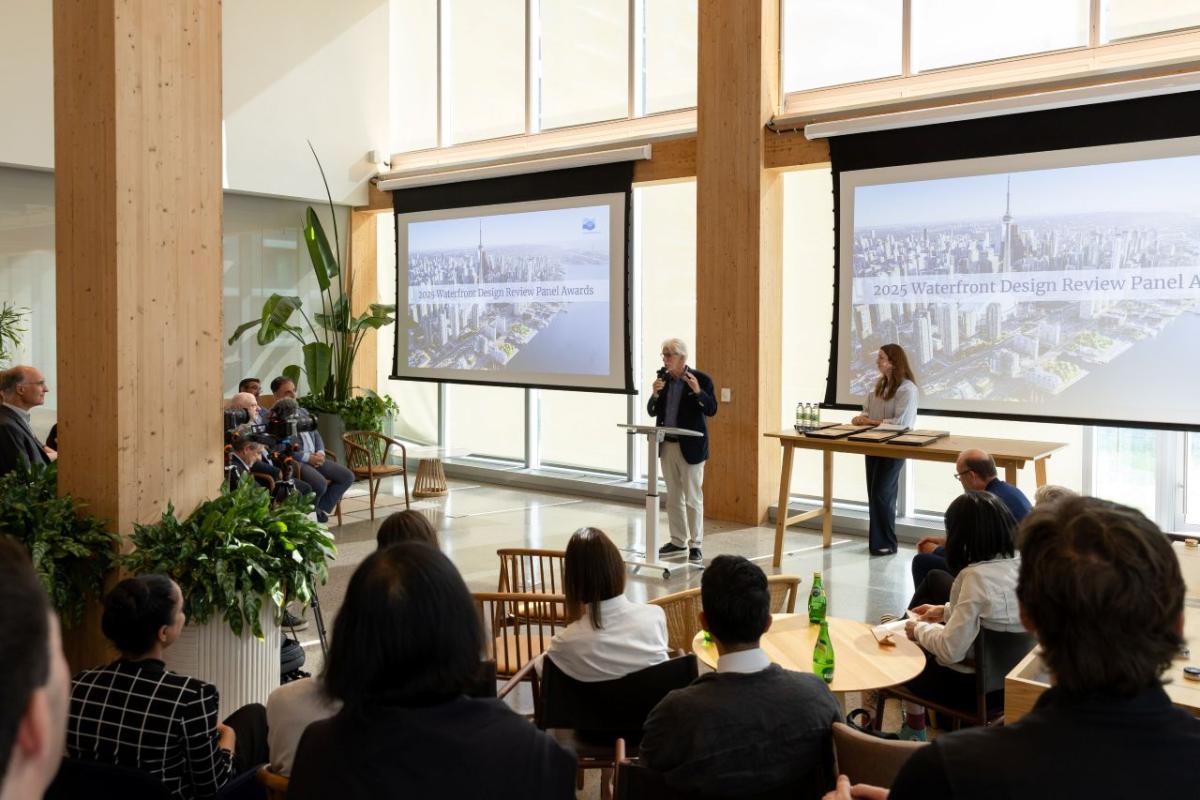 A person stands at a podium speaking to an audience in a modern conference room. Two large screens display a "2025 Waterfront Design Review Panel Awards" presentation, featuring an aerial cityscape image. Several people are seated, listening attentively.