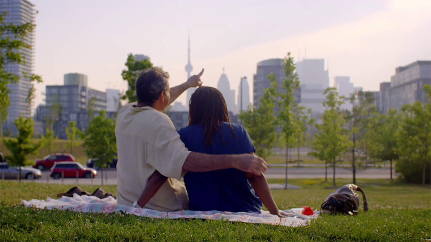 A man and a woman sit on a grassy hill, looking over the Toronto skyline