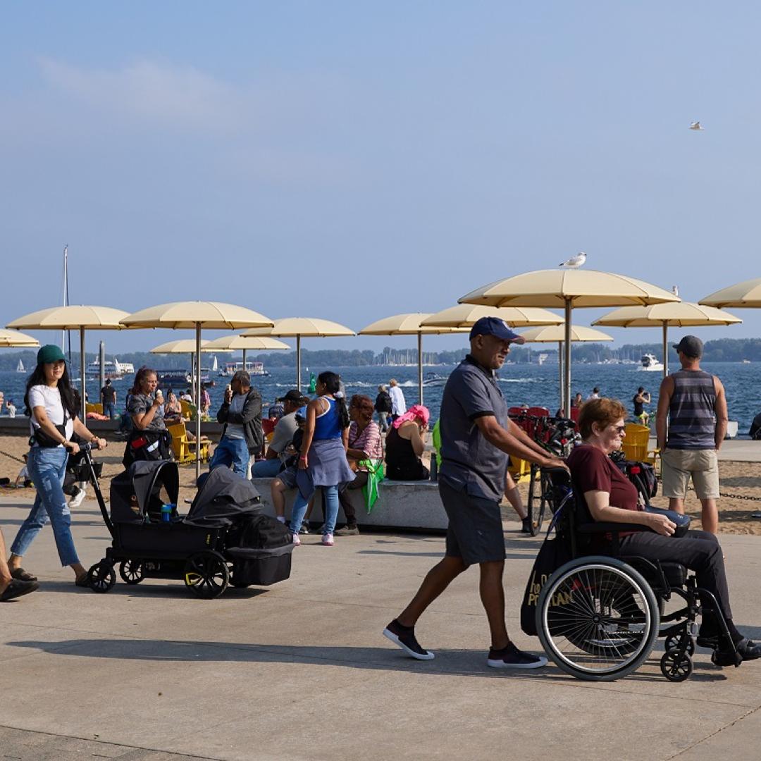 People at an urban beach on a summer day. One person is using a wheelchair, another is pushing a stroller.