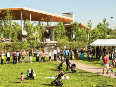 People standing and sitting on a grassy lawn in a park. A pavilion in the background. 