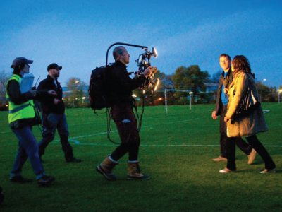 A camera crew filming two people walking on grass at sunrise.