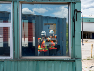 Two photographers in construction safety equipment taking a picture of themselves in a window.