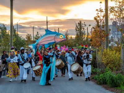 A large group of people participate in an outdoor parade at sunset along a planted pathway at Biidaasige Park. At the front of the procession, several drummers walk in a line, and a performer holds a large, colourful fish puppet. Participants behind them carry additional bright, handmade puppets and decorations. 