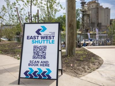 A sign board on a street that reads: East West Shuttle, scan and book here.