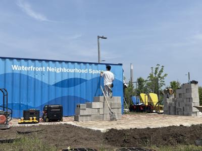 A person stencils on the words "Waterfront Neighbourhood Space" on to the side of a blue painted shipping container