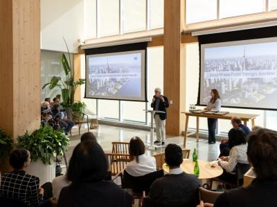 A person stands at a podium speaking to an audience in a modern conference room. Two large screens display a "2025 Waterfront Design Review Panel Awards" presentation, featuring an aerial cityscape image. Several people are seated, listening attentively.