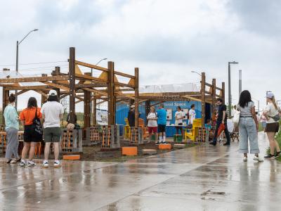 Visitors gather at an outdoor community space on a rainy day. Several people stand and walk along a wet concrete pathway beside a partially constructed wooden pavilion. A blue shipping container with informational displays serves as a backdrop, while chairs, tables, and construction materials are arranged throughout the site. Overcast skies and reflective pavement emphasize the rainy weather.