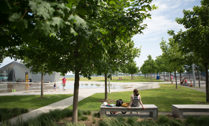 people sitting on a park bench next to some trees and a splash pad