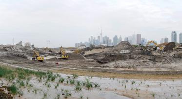 Excavation of the future river valley in the Port Lands. In the foreground, small plants are growing out of a muddy area. In the midground, heavy machinery is working on large piles of soil. The Toronto Skyline is in the background.