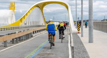 Cyclists ride in a separated bike lane through a yellow and white arched bridge.