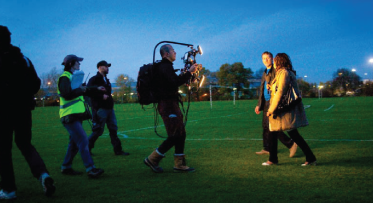 A camera crew filming two people walking on grass at sunrise.