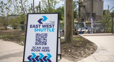 A sign board on a street that reads: East West Shuttle, scan and book here.