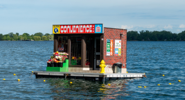 A floating convenience store artwork stocked with flowers and everyday items sits on the water in daylight, featuring a bright “Convenience” sign, an “ATM” sign, and a yellow fire hydrant on the platform.