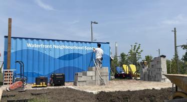 A person stencils on the words "Waterfront Neighbourhood Space" on to the side of a blue painted shipping container
