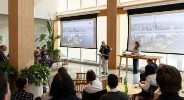 A person stands at a podium speaking to an audience in a modern conference room. Two large screens display a "2025 Waterfront Design Review Panel Awards" presentation, featuring an aerial cityscape image. Several people are seated, listening attentively.