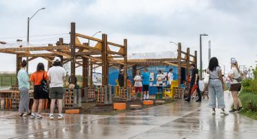 Visitors gather at an outdoor community space on a rainy day. Several people stand and walk along a wet concrete pathway beside a partially constructed wooden pavilion. A blue shipping container with informational displays serves as a backdrop, while chairs, tables, and construction materials are arranged throughout the site. Overcast skies and reflective pavement emphasize the rainy weather.